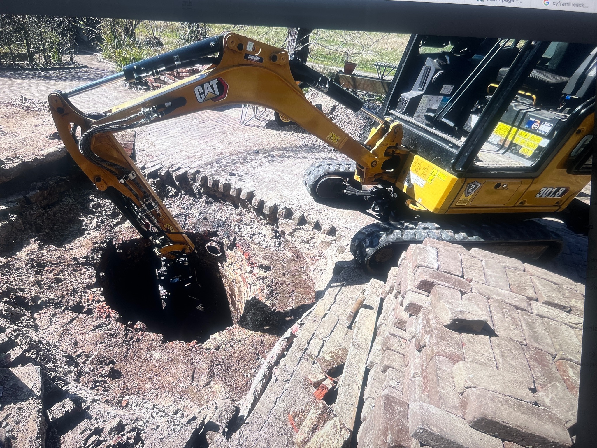 Żółta minikoparka CAT 301.8 w trakcie wykopu okrągłego otworu w ziemi, obok stos kostki brukowej. Prace ziemne na posesji.