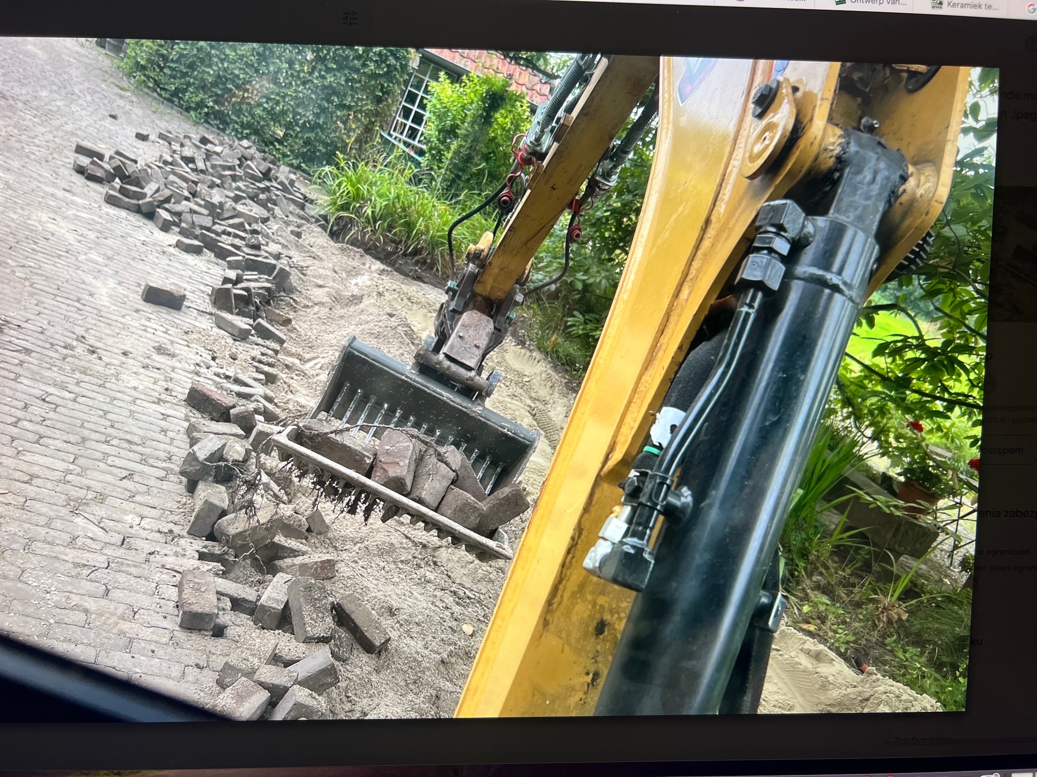 Żółta koparka z łyżką pełną kostki brukowej na piasku, w tle zielona roślinność i fragment budynku. Prace ziemne w toku, przygotowanie podłoża pod nową nawierzchnię.