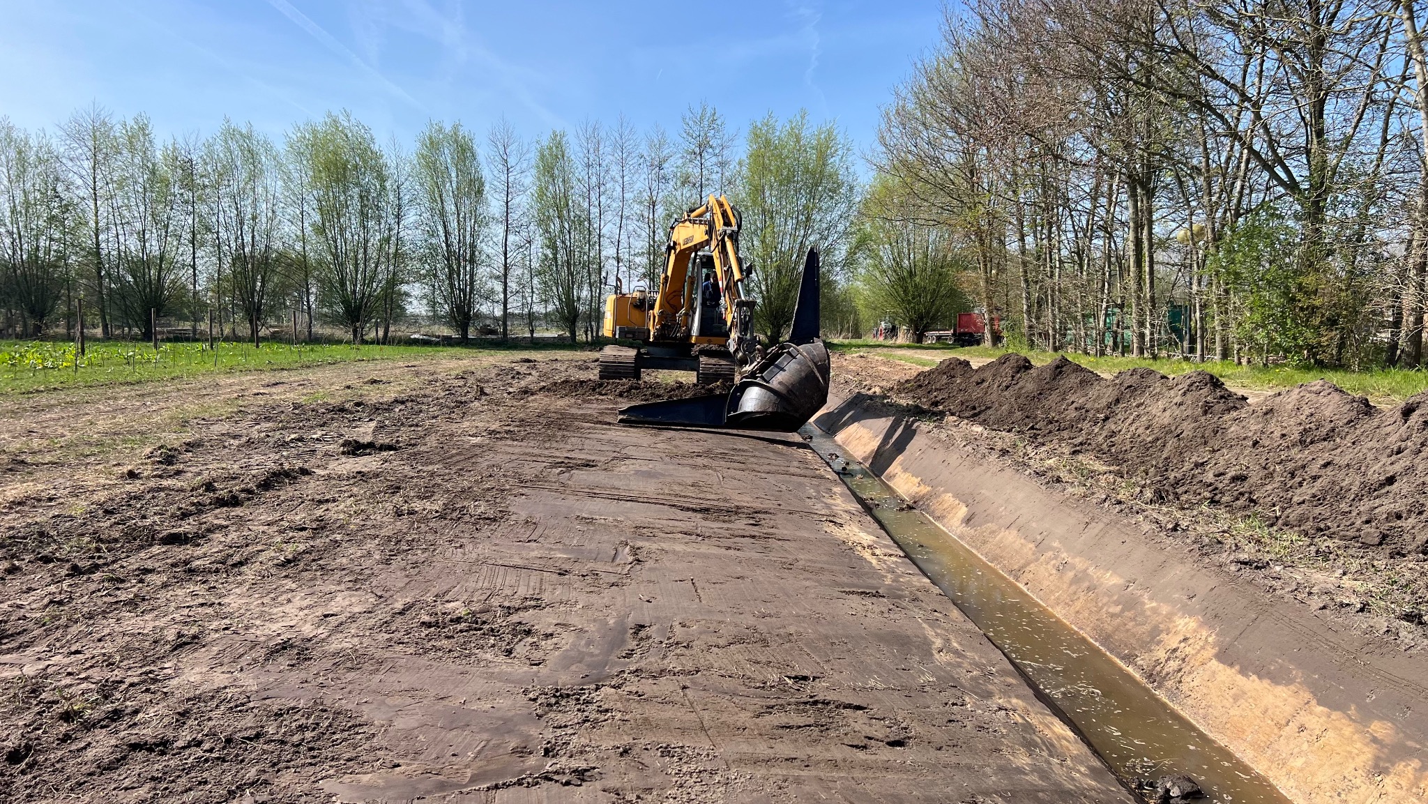 Żółta koparka rowowa w trakcie pogłębiania rowu melioracyjnego na polu. W tle drzewa i błękitne niebo. Widoczne świeżo wykopane ziemia i woda w rowie.