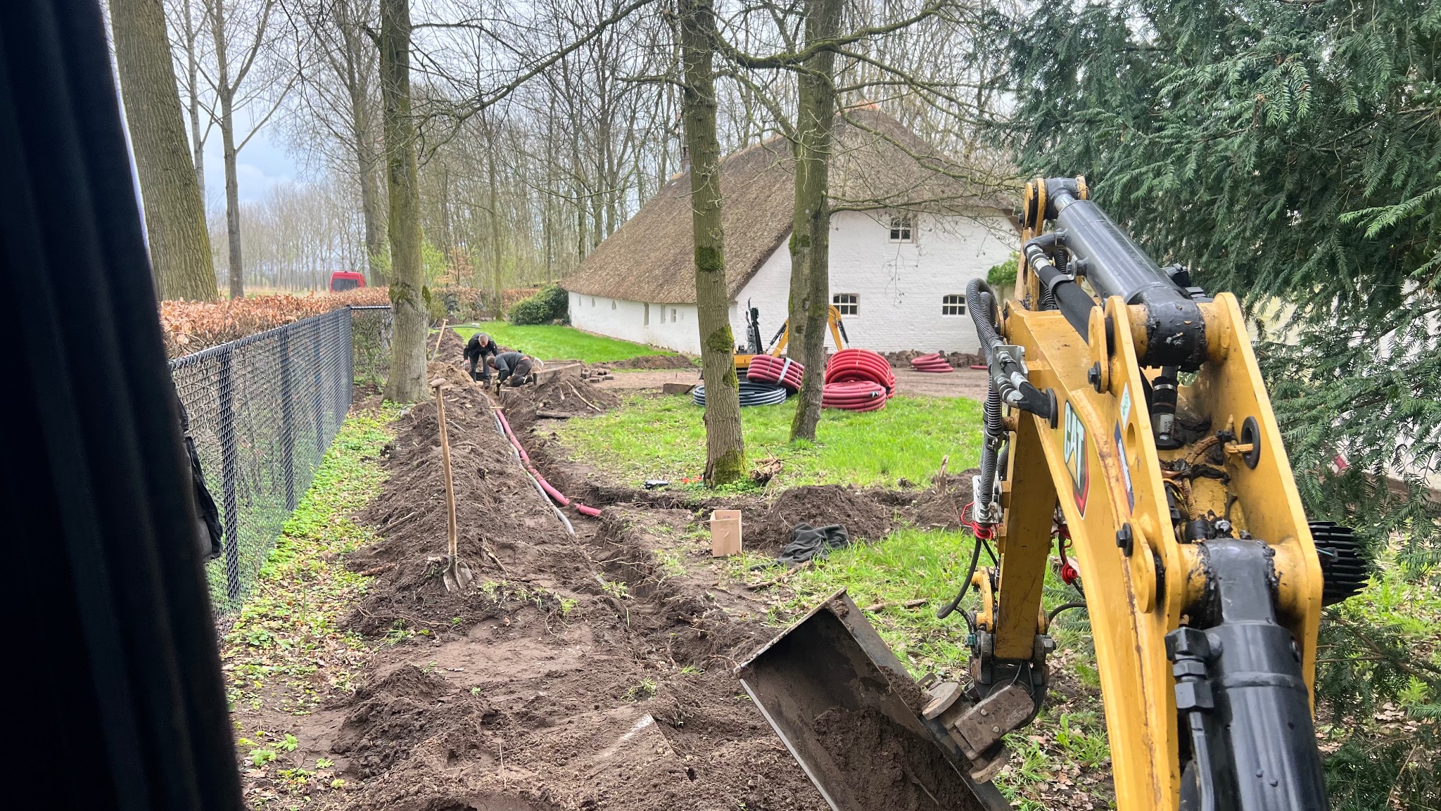 Wykop pod instalacje: żółta koparka w akcji, widoczne rury i pracownicy w tle, dom z strzechą. Prace ziemne w toku, teren przygotowywany pod ułożenie instalacji.