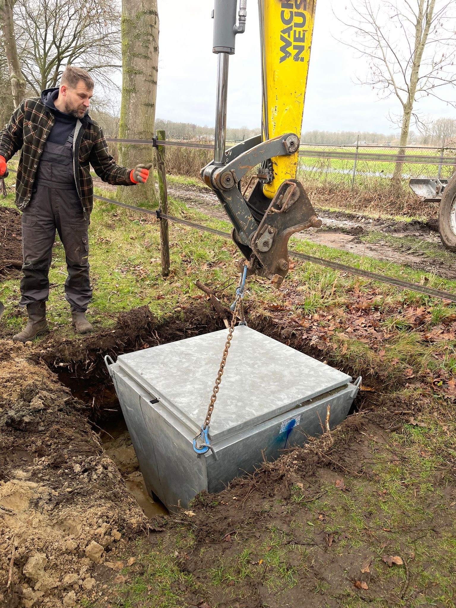 Montaż szamba z użyciem koparki: pracownik w kombinezonie nadzoruje opuszczanie metalowego zbiornika do wykopu. Widoczna maszyna Wacker Neuson i błotniste otoczenie.
