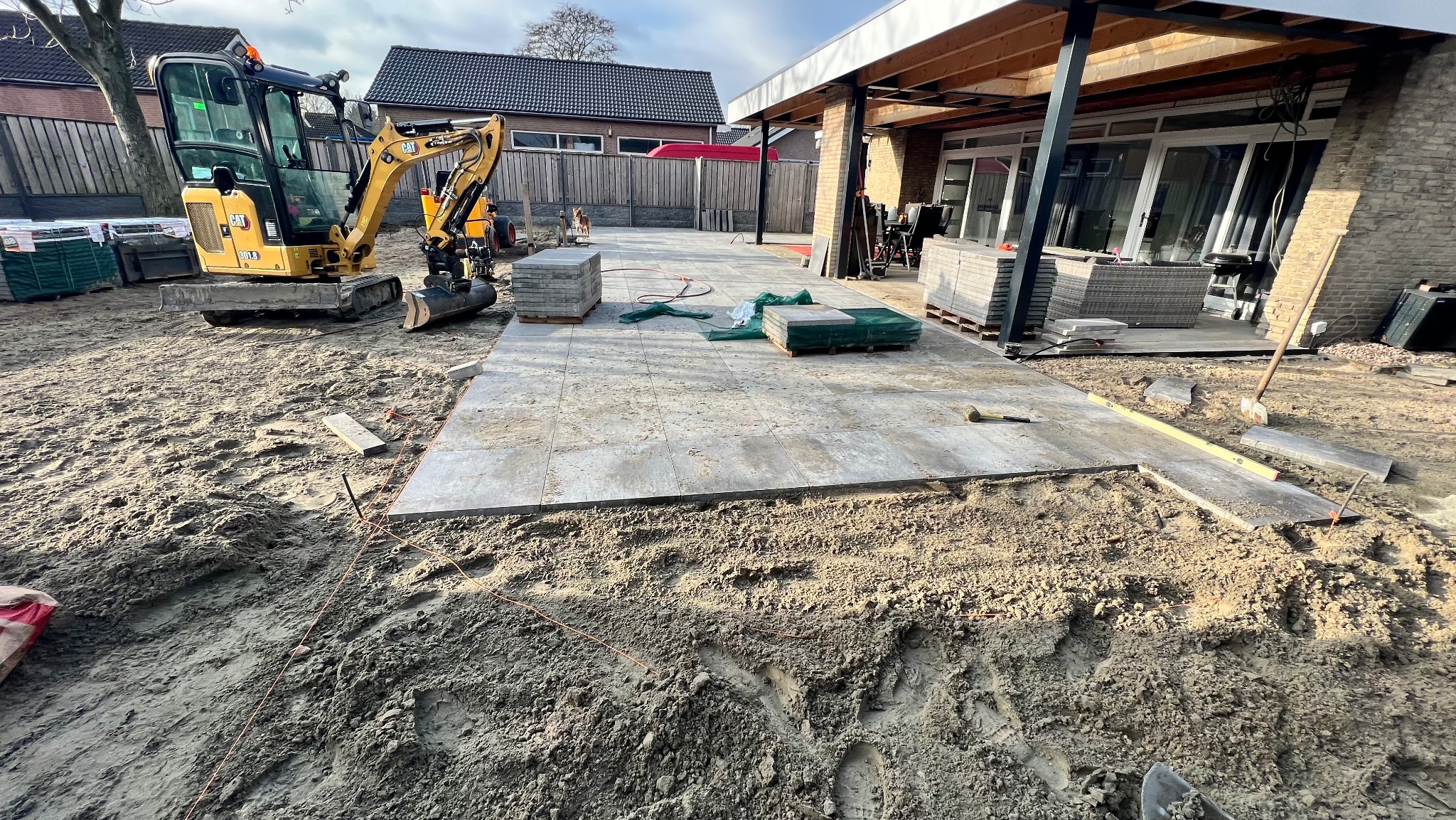 Układanie szarych płyt na tarasie w ogrodzie. Mini koparka CAT i palety z kostką w tle. Częściowo ułożona nawierzchnia, widoczne narzędzia i poziomica.