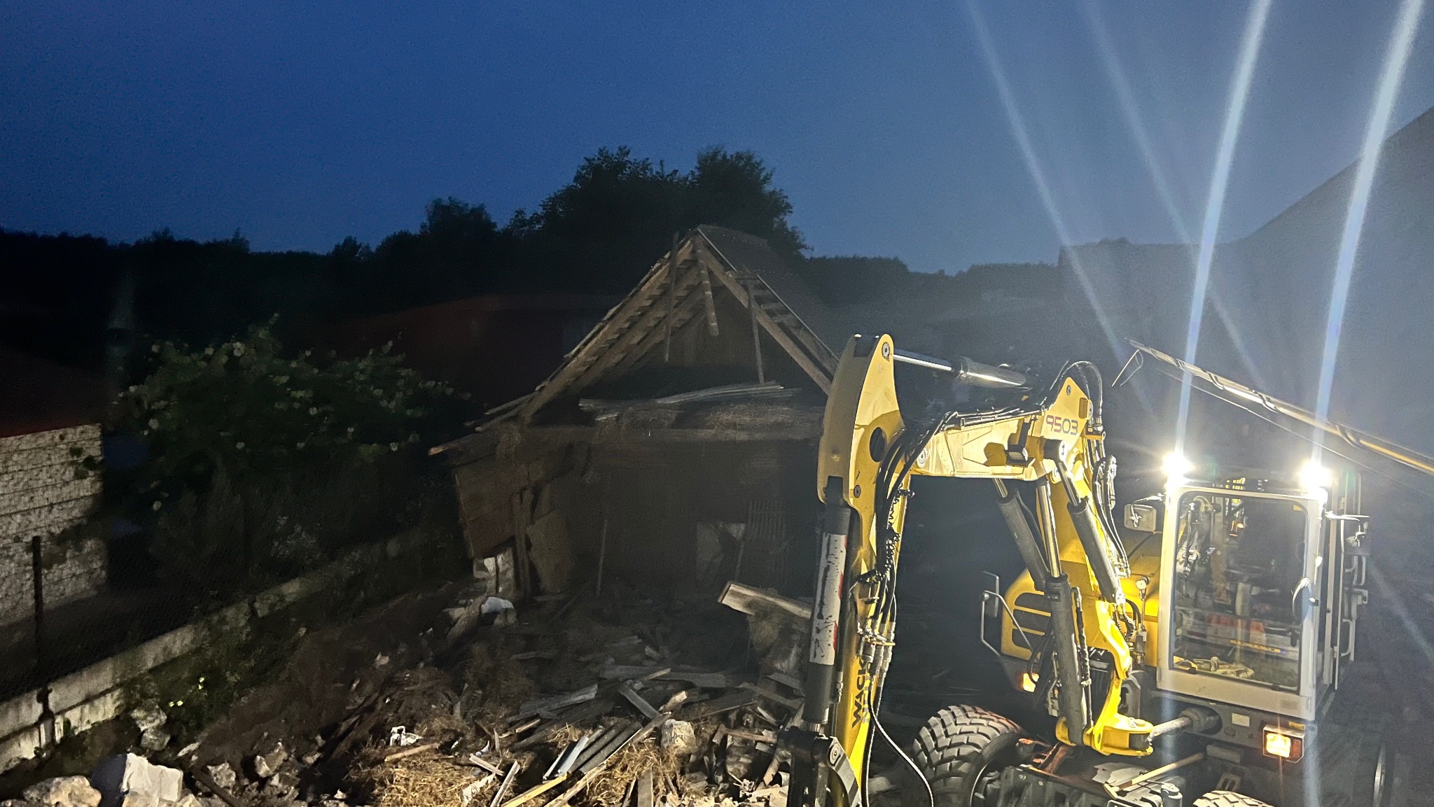 Żółta koparka przy rozbiórce starego, drewnianego budynku w nocy. Widoczne resztki konstrukcji i gruz. Oświetlenie koparki rozjaśnia ciemne otoczenie.