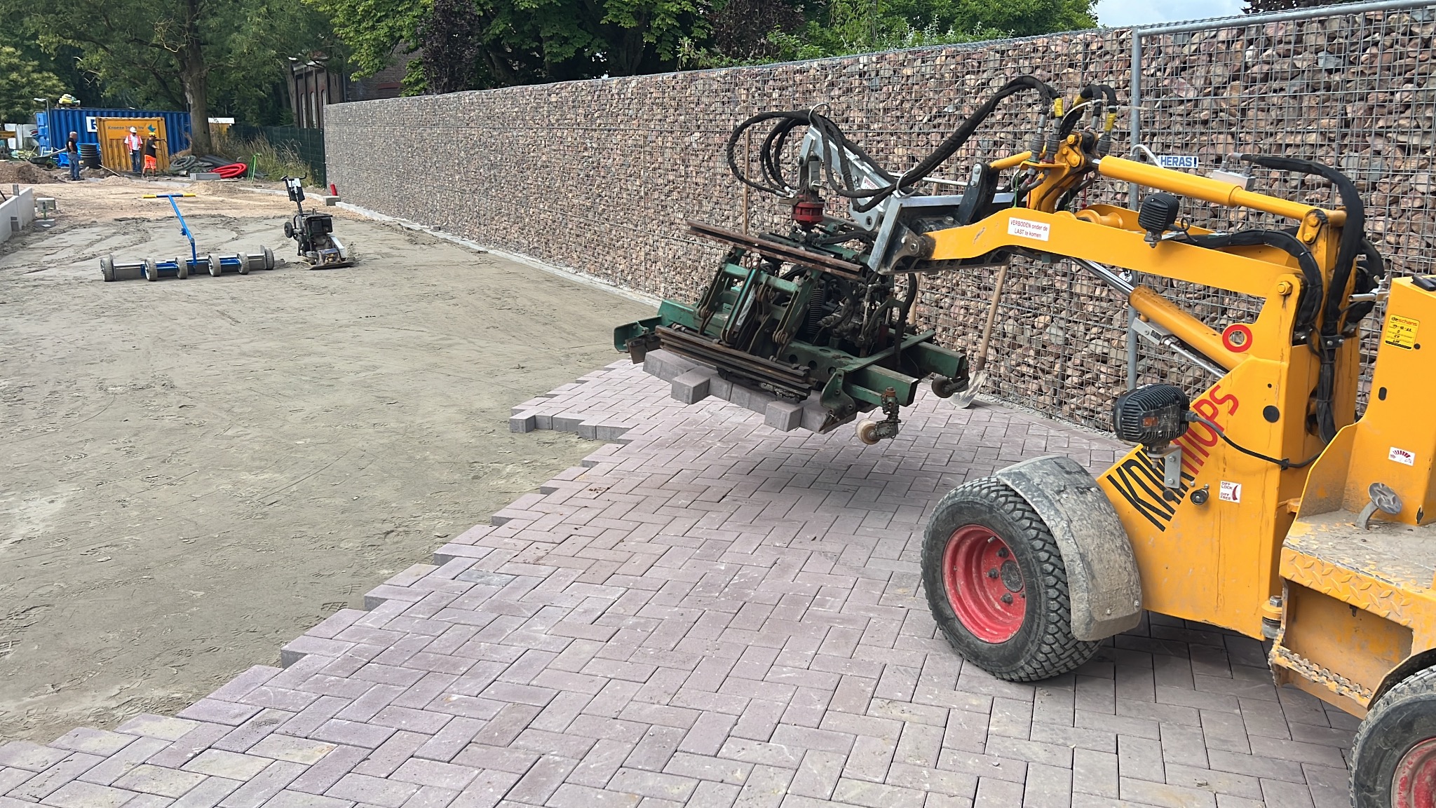 Układanie kostki brukowej maszyną. Żółty podnośnik z zielonym chwytakiem układa kostkę w wzór jodełki. W tle przygotowany teren i mur z kamieni.