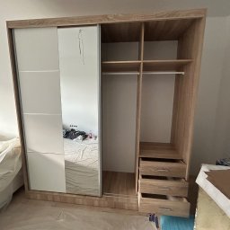 Maksym Jastrebov - Szafa z przesuwnymi drzwiami, częściowo zamontowana, z widocznymi półkami i szufladami. W tle łóżko przykryte folią ochronną, co sugeruje trwający remont.