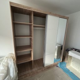 Maksym Jastrebov - Jasny, drewniany regał z przesuwnymi drzwiami, częściowo rozpakowany z folii ochronnej, stoi na podłodze zabezpieczonej folią malarską.