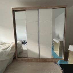 Maksym Jastrebov - Jasna szafa przesuwna z lustrami, częściowo zasłonięta folią ochronną, w trakcie montażu. Widoczne elementy wyposażenia wnętrza w tle. Ujęcie z bliska.