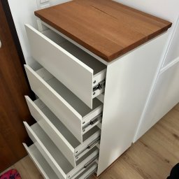 Maksym Jastrebov - Biała komoda z drewnianym blatem i wysuniętymi szufladami, ukazująca mechanizmy. Ujęcie z góry, widoczna podłoga i fragment ściany z włącznikami.