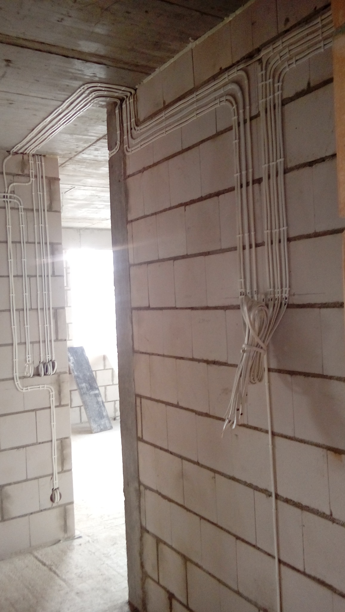 Białe kable elektryczne ułożone w peszlach na ścianie z bloczków w surowym wnętrzu budynku w trakcie budowy. Widoczne załamania kabli pod kątem prostym.