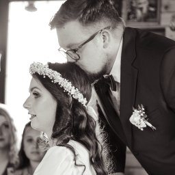 ALTITUDE ATTITUDE - Aleksandra Słojewska - Czarno-białe zdjęcie: Pan młody w garniturze całuje w głowę pannę młodą z wiankiem na włosach. W tle goście i jasne okno.