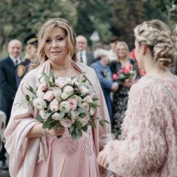 ALTITUDE ATTITUDE - Aleksandra Słojewska - Panna młoda w pudrowej sukni z bukietem, otoczona gośćmi weselnymi. Ujęcie z lekkiej perspektywy, miękkie światło podkreśla detale sukni i fryzury.