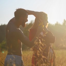 ALTITUDE ATTITUDE - Aleksandra Słojewska - Para w kontraście słońca na łące: mężczyzna bez koszuli delikatnie dotyka włosów kobiety trzymającej żółty kwiat. Romantyczna atmosfera, naturalne światło.