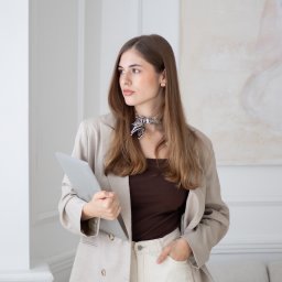 ALTITUDE ATTITUDE - Aleksandra Słojewska - Portret kobiety w beżowym garniturze, trzymającej laptopa, Pruszków. Ujęcie w neutralnym wnętrzu, z delikatnym makijażem i apaszką. Profesjonalny wizerunek.