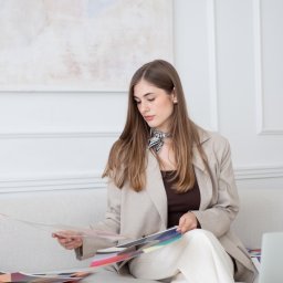 ALTITUDE ATTITUDE - Aleksandra Słojewska - Kobieta w beżowym żakiecie siedzi na kanapie, przeglądając paletę kolorów. Na stoliku leży laptop. W tle widoczny obraz na ścianie i dekoracyjne panele.