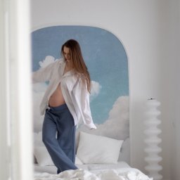 Aleksandra Słojewska - Kobieta w ciąży, w rozpiętej białej koszuli i dżinsach, stoi na łóżku na tle ściany z motywem nieba. Ujęcie z dołu, widoczny brzuch i długie włosy.