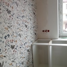 Elżbieta Wierzbicka - Ściana z tapetą w kwiaty w kuchni, widoczne białe szafki bez blatów i okno. Gniazdka elektryczne na ścianie. Ujęcie z dołu, podkreślające wzór tapety.