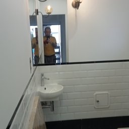 Elżbieta Wierzbicka - Wykończona łazienka z białą armaturą, lustrem i oświetleniem. Na ścianach białe płytki typu metro z czarnym pasem. Na podłodze wzorzysta mozaika. Widoczne odbicie osoby w lustrze.