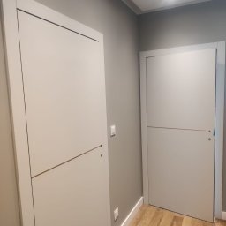 Elżbieta Wierzbicka - Dwa nowoczesne, białe drzwi wewnętrzne z poziomymi akcentami w szarej ścianie, widoczne gniazdko i włącznik, drewnopodobna podłoga. Minimalistyczny design.