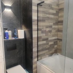 Elżbieta Wierzbicka - Nowoczesna łazienka po remoncie: ciemne płytki imitujące marmur, drewnopodobne panele na ścianie prysznica, biała wanna z parawanem i toaleta. Oświetlenie punktowe.