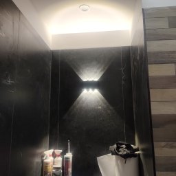 Elżbieta Wierzbicka - Nowoczesna łazienka z czarnymi płytkami imitującymi marmur, oświetleniem punktowym i półką z akcesoriami. Widoczne detale wykończenia i materiały budowlane.