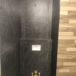 Elżbieta Wierzbicka - Częściowo wykończona łazienka z czarnymi płytkami imitującymi marmur i drewnopodobnymi płytkami na ścianie. Widoczna instalacja podtynkowa WC i podejścia wodne.