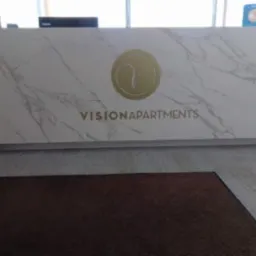 Biała lada recepcyjna z marmurowym wzorem i złotym logo 'VISIONAPARTMENTS'. Nowoczesny design, widok z przodu, dywan w odcieniach brązu na podłodze.