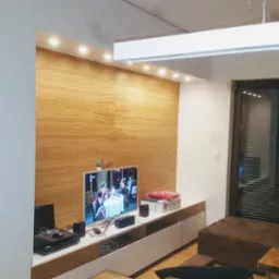 Salon z drewnianą ścianą, podświetleniem LED, telewizorem i nowoczesnymi meblami, widoczne również przesuwne drzwi balkonowe.