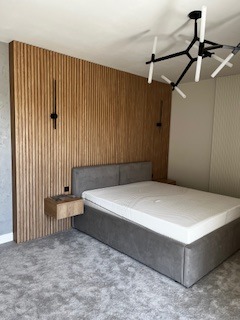 Sypialnia z łóżkiem kontynentalnym w szarej tapicerce, drewnianym panelem ściennym z pionowych listew, minimalistyczną lampą wiszącą i szafką nocną w kolorze drewna.