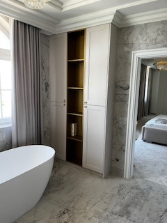 Jasna łazienka z wolnostojącą wanną, marmurową podłogą i wysoką, wąską szafą z półkami w kolorze złota, widok na sypialnię przez otwarte drzwi.