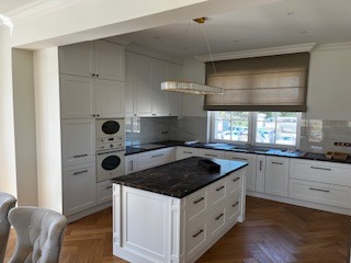 Jasna kuchnia z wyspą i parkietem w jodełkę, oświetlona nowoczesną lampą wiszącą, widok z perspektywy lekko z boku.