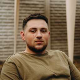 Frilans - Portret młodego mężczyzny z brodą w zielonej koszulce na tle drewnianego płotu i jasnej konstrukcji. Ujęcie z bliska, naturalne światło.