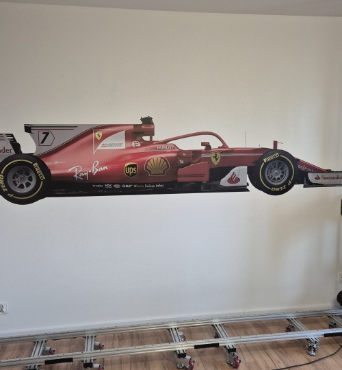 Wielkoformatowy wydruk bolidu Formuły 1 Ferrari na ścianie, widoczny fragment toru jezdnego kamery. Realistyczne odwzorowanie detali, matowe wykończenie.