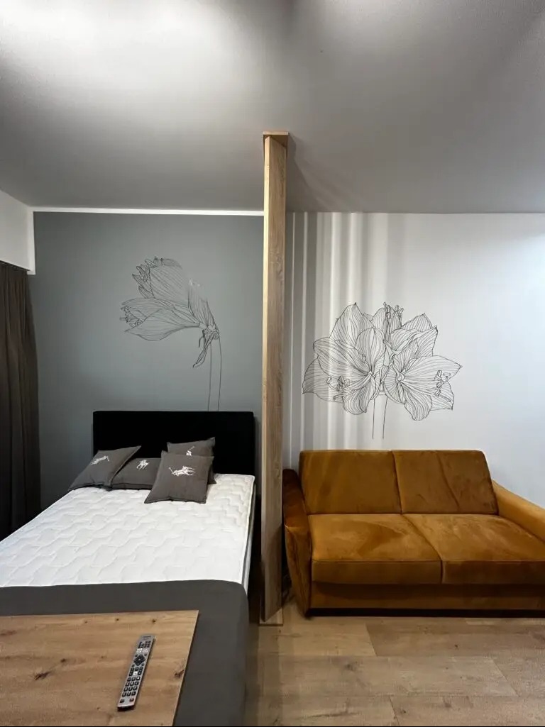 Pokój podzielony drewnianą przegrodą: z jednej strony łóżko z szarą pościelą, z drugiej sofa w odcieniach brązu. Na ścianach rysunki kwiatów.