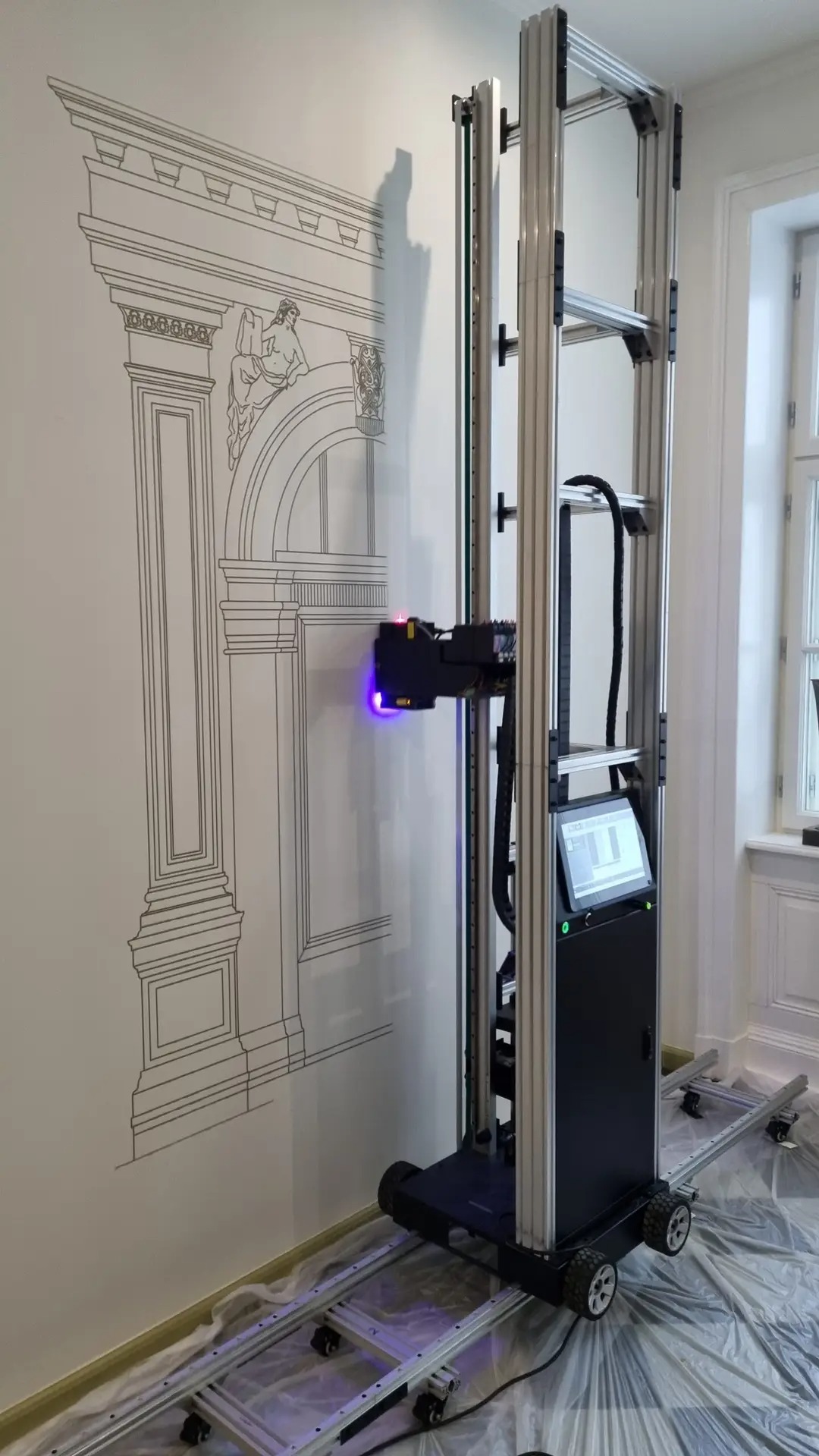 Drukarka pionowa w trakcie nanoszenia czarnego konturu klasycznej architektury na białą ścianę. Widoczne prowadnice i ekran urządzenia.