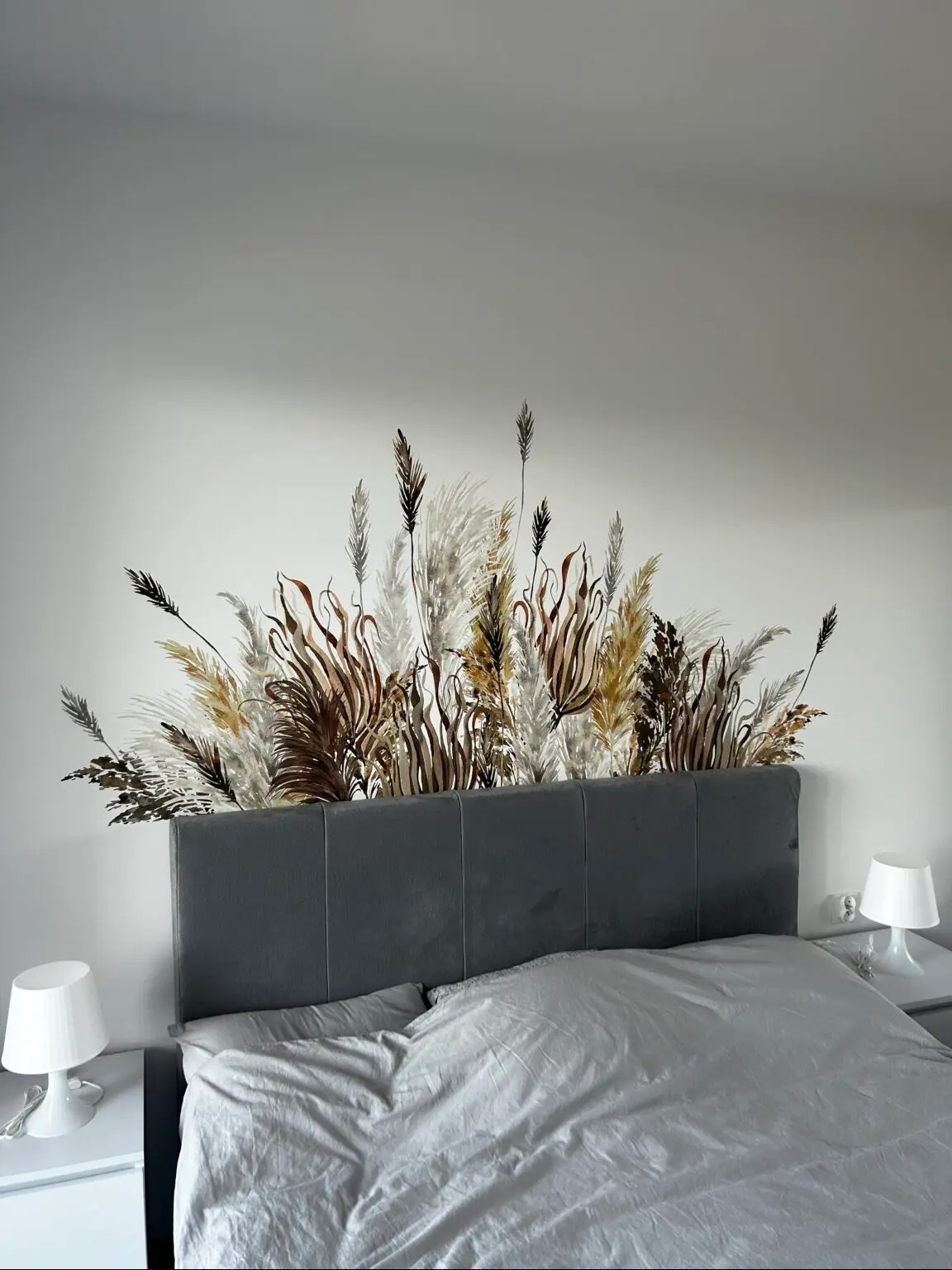 Naklejka dekoracyjna z motywem traw ozdobnych nad wezgłowiem łóżka w sypialni. Realizacja grafiki na ścianie w neutralnej kolorystyce.