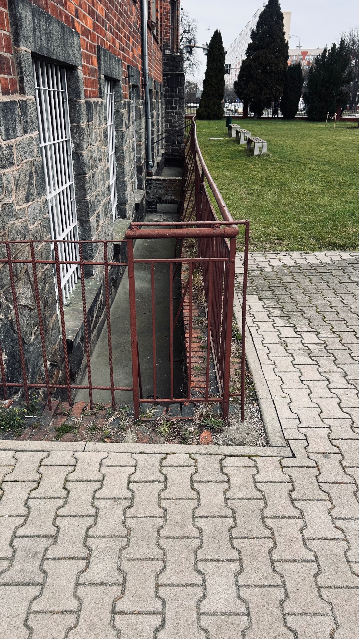 Metalowa barierka zabezpieczająca zejście do piwnicy przy budynku z cegły i kamienia. Widoczne okna piwniczne z kratami i chodnik z kostki.
