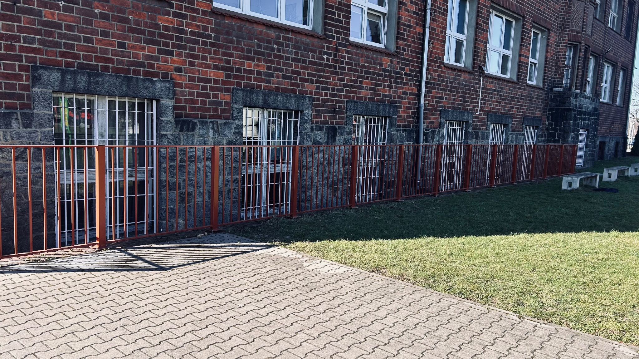 Czerwone, metalowe ogrodzenie okienne i obok budynku z cegły, z widoczną kostką brukową i trawnikiem. Ochrona okien piwnicznych.