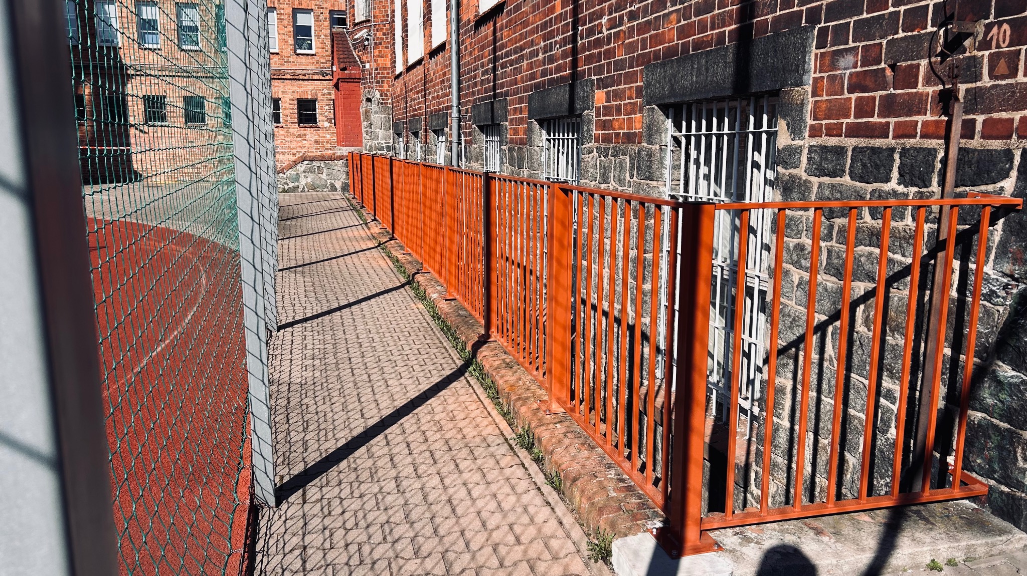 Pomarańczowa balustrada metalowa przy chodniku z kostki, w tle budynek z cegły i siatka ogrodzeniowa boiska. Widok z boku, oświetlenie słoneczne.