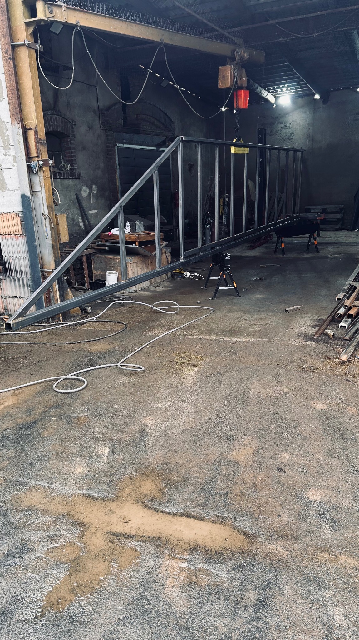 Surowa, metalowa konstrukcja balustrady podwieszona w warsztacie. Widoczne narzędzia, surowe ściany i betonowa podłoga z plamami. Ujęcie z niskiej perspektywy.