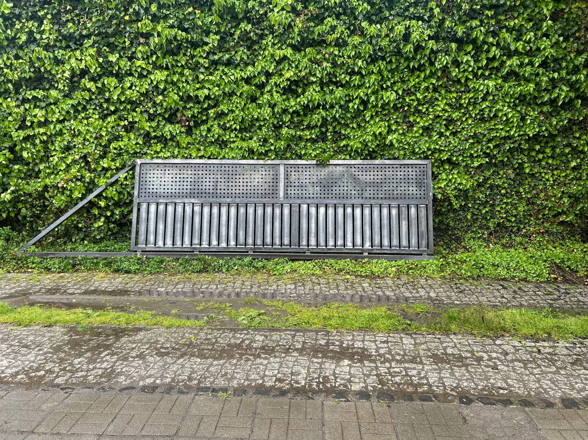 Metalowa brama segmentowa z perforowaną górną częścią, leżąca na mokrym bruku przed ścianą z bluszczu. Widoczny fragment ramy i mechanizmu bramy.
