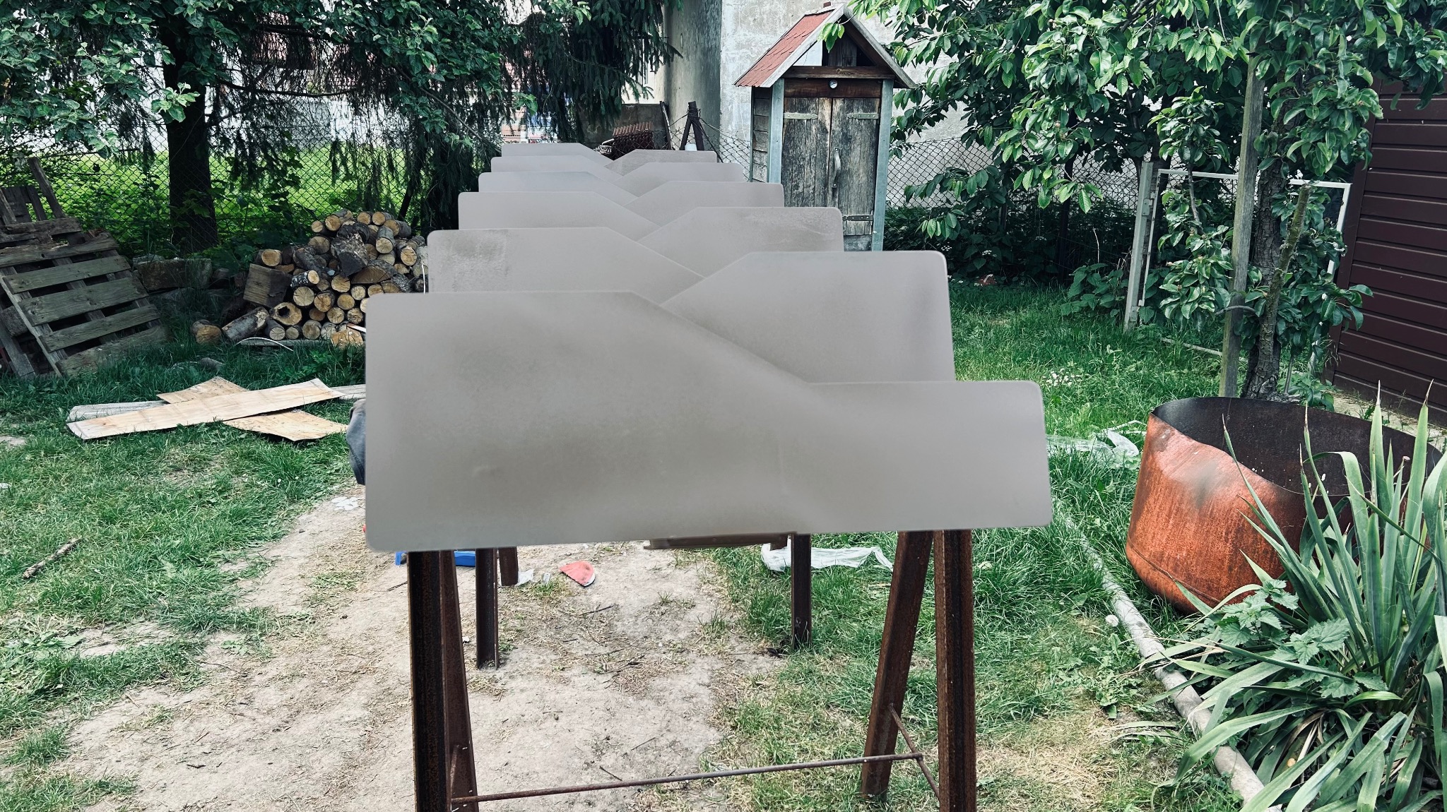 Przygotowane elementy metalowe, prawdopodobnie do ogrodzenia, ustawione na kozłach w ogrodzie, czekające na dalszą obróbkę lub montaż. Widoczne ślady farby.