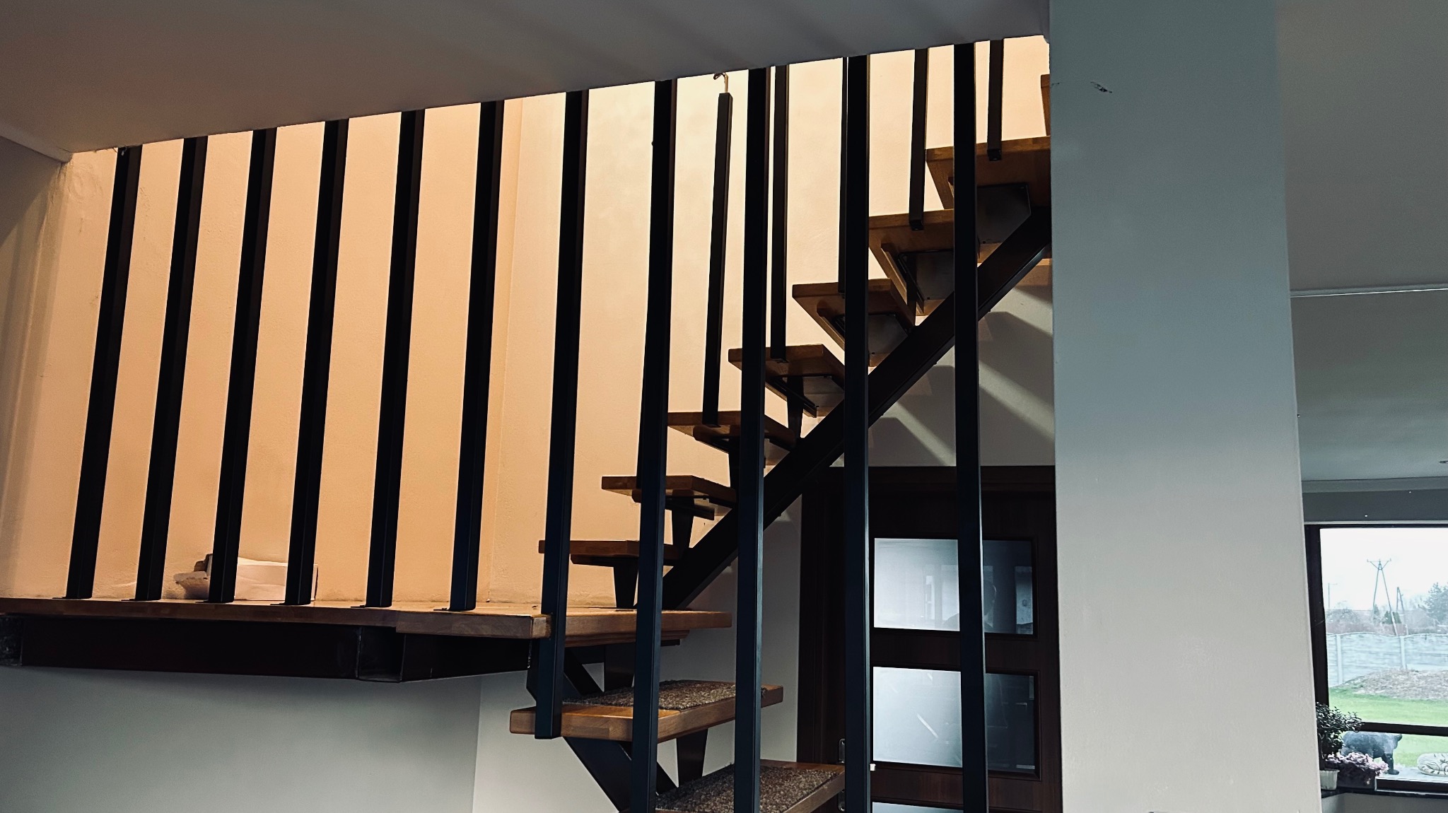 Nowoczesne schody metalowe z drewnianymi stopniami i czarnymi, pionowymi balustradami, widoczne z dołu, z oknem w tle. Minimalistyczny design.