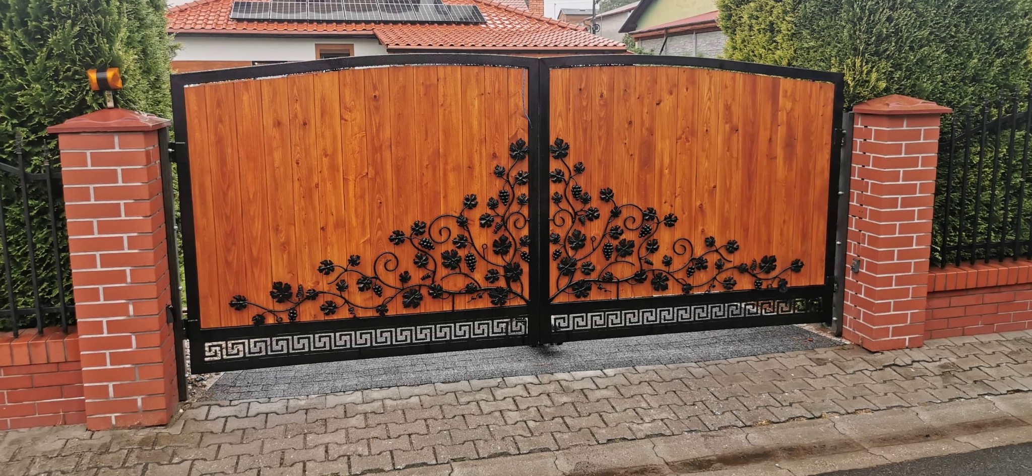 Dwuskrzydłowa brama wjazdowa z drewnianym wypełnieniem i metalową dekoracją z motywem winorośli, osadzona między murowanymi słupkami.