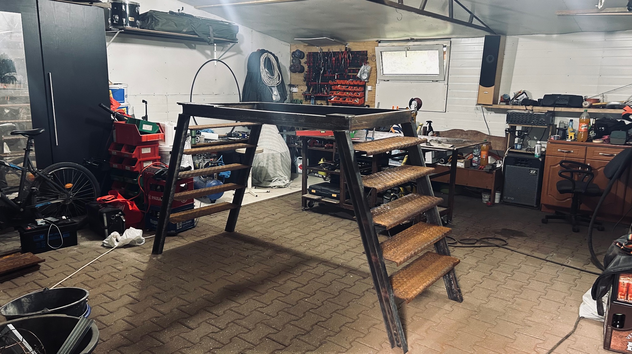 Metalowa konstrukcja schodów z platformą, wykonana w warsztacie w Kunicach. Widoczne spawy i surowy metal, otoczenie warsztatowe z narzędziami i akcesoriami.