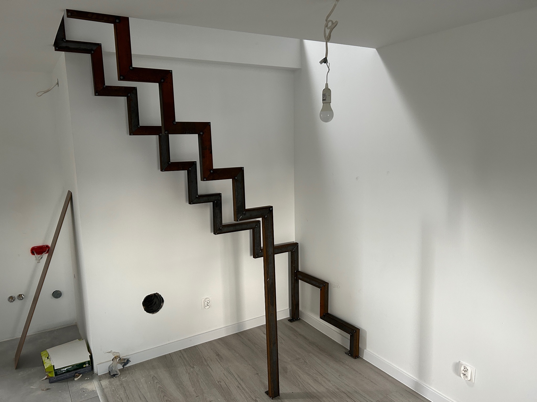 Surowe, metalowe schody o zygzakowatym kształcie, jeszcze bez wykończenia, zamontowane w jasnym wnętrzu. Widoczne ślady prac montażowych na ścianach.
