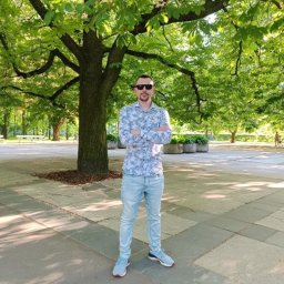 Sławomir Skoworodko - Mężczyzna w okularach przeciwsłonecznych stoi na chodniku z szarej kostki, w tle drzewo i park. Ubrany w jasne jeansy i koszulę w drobny wzór.