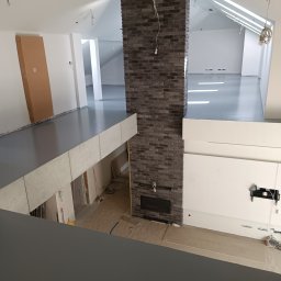 Construction World - Wnętrze domu w trakcie wykańczania: szara posadzka żywiczna na antresoli, ceglany komin, surowe ściany i podłogi na dole. Widok z góry na dwupoziomowe pomieszczenie.
