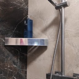 Bogusław Pytel BP Company - Nowoczesna kabina prysznicowa z chromowaną armaturą i półką, na której stoi butelka kosmetyku. Ściany wyłożone płytkami imitującymi marmur w odcieniach szarości.