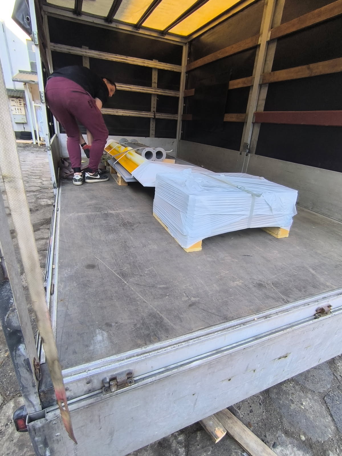 Załadunek elementów konstrukcyjnych, prawdopodobnie stalowych, do wnętrza samochodu dostawczego. Widoczne arkusze blachy i rury na palecie, osoba wewnątrz pojazdu.