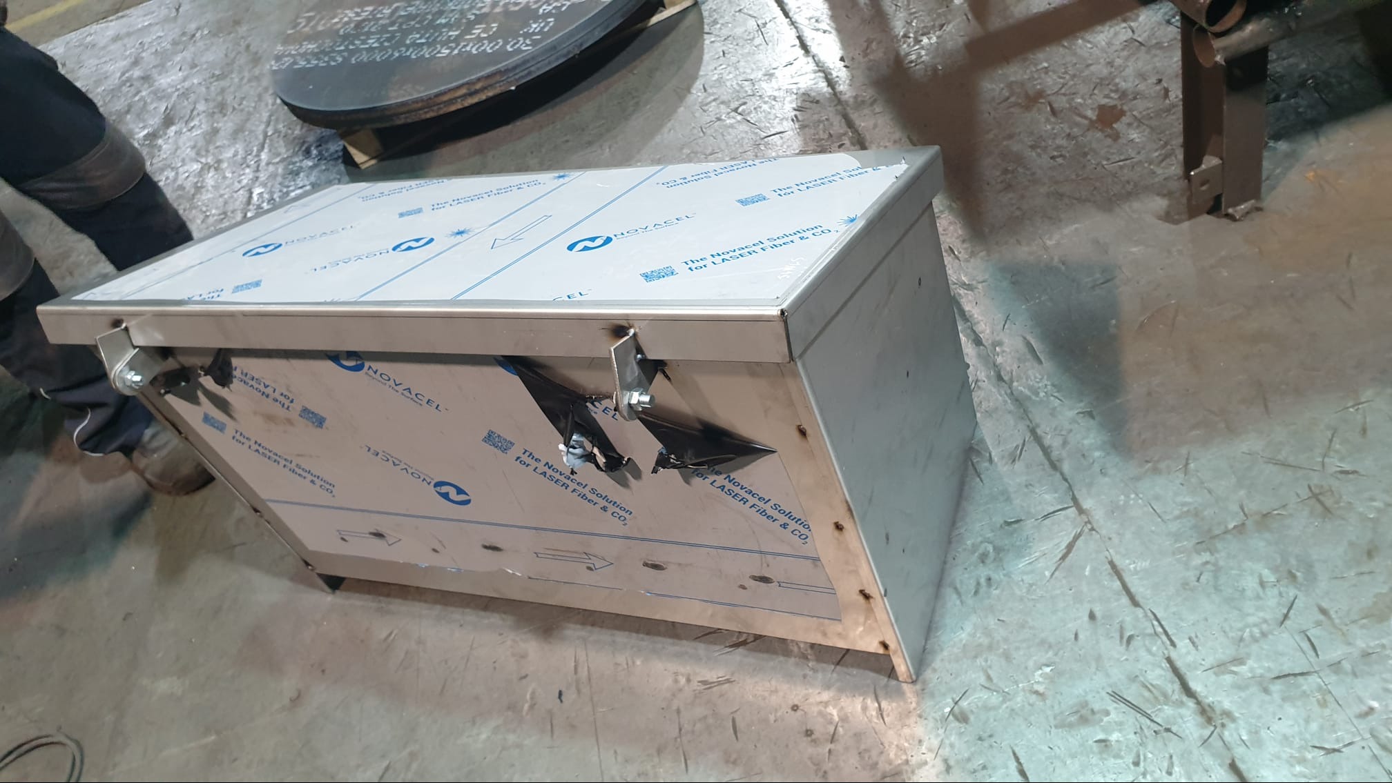 Metalowa konstrukcja z elementami mocującymi, pokryta folią ochronną Novacel, w warsztacie. Widoczne ślady obróbki i niedoskonałości powierzchni.